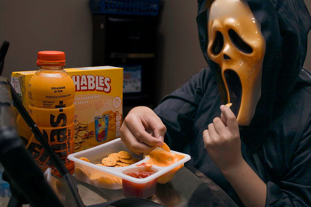 Person in a scream mask and hooded robe eating from a snack tray with crackers and dips, alongside a Lunchables box and a drink bottle.