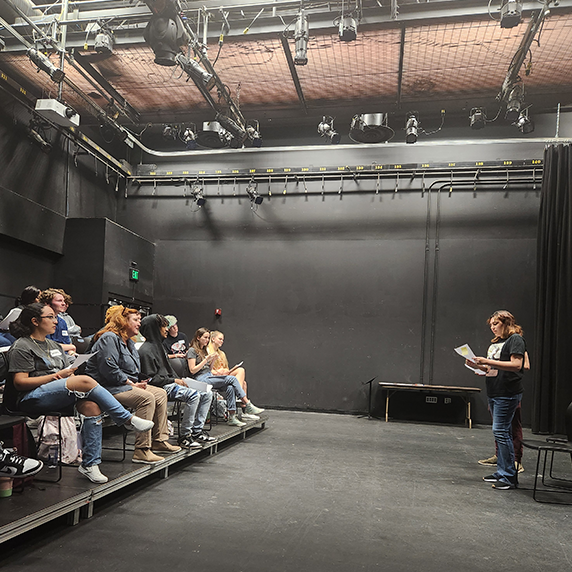 A theater space with seated audience on the left and a standing person holding a script on the right.