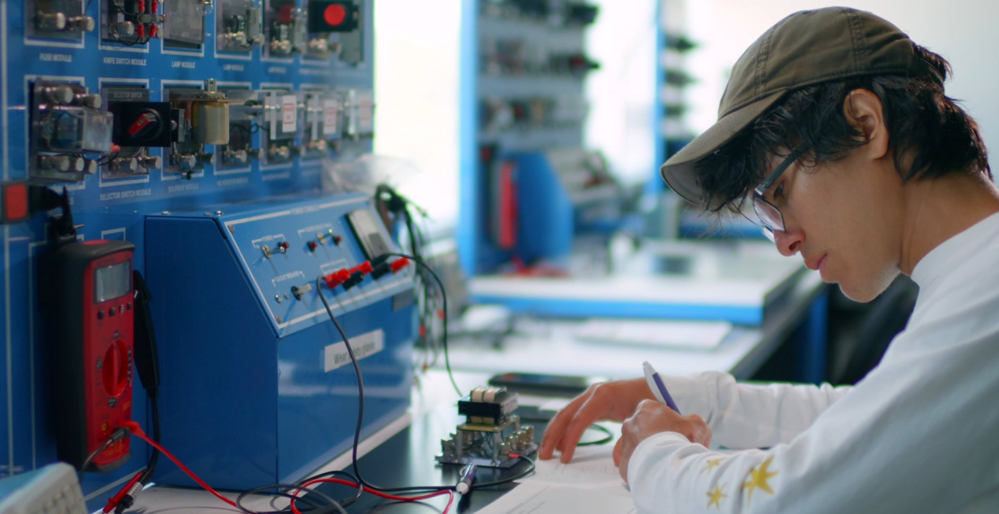 A person writing at a table with electronic equipment and a blue control panel in a laboratory.