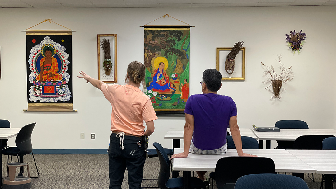 Two people in a room observe artworks, including paintings and framed masks, on a wall.