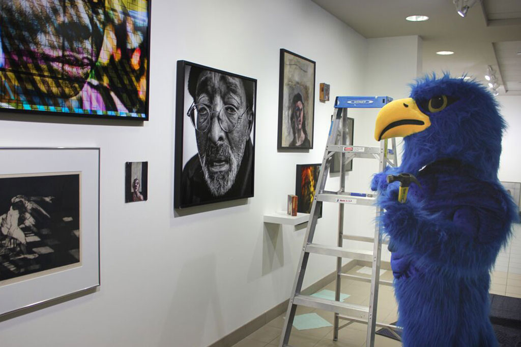 HCC Hawk mascot in an art gallery with various artworks on the wall and a step ladder nearby.