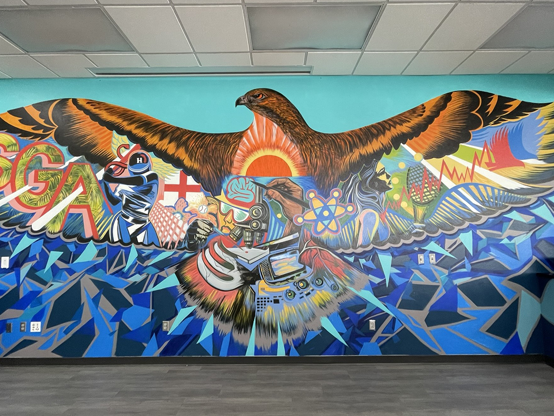 A vibrant mural of an eagle with outstretched wings, surrounded by initials, symbols, and geometric patterns on a blue background.