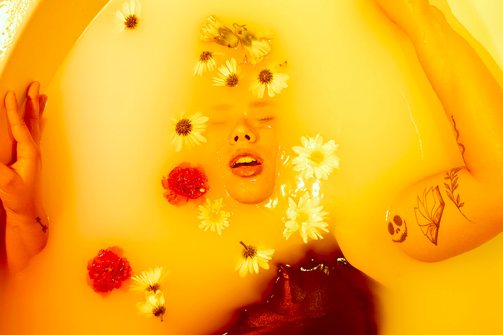 A person submerged in bright yellow water with flowers floating around their face, eyes closed, and tattoos visible on the arm.