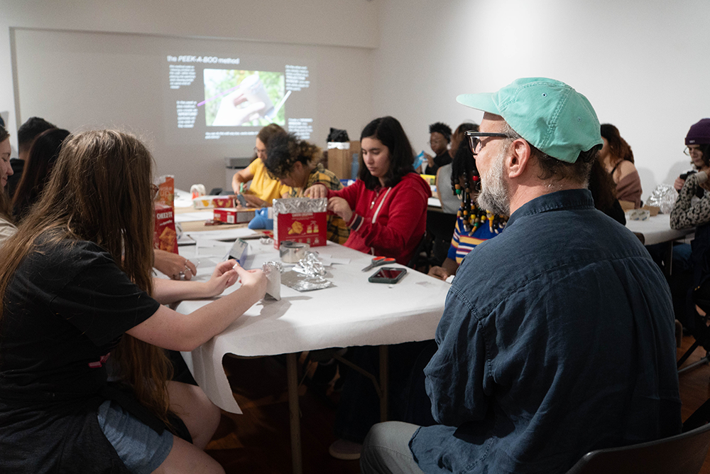 A group of people at a table in a workshop setting, engaged in a crafting activity with a presentation on the "PEEK-A-BOO method" projected in the background.