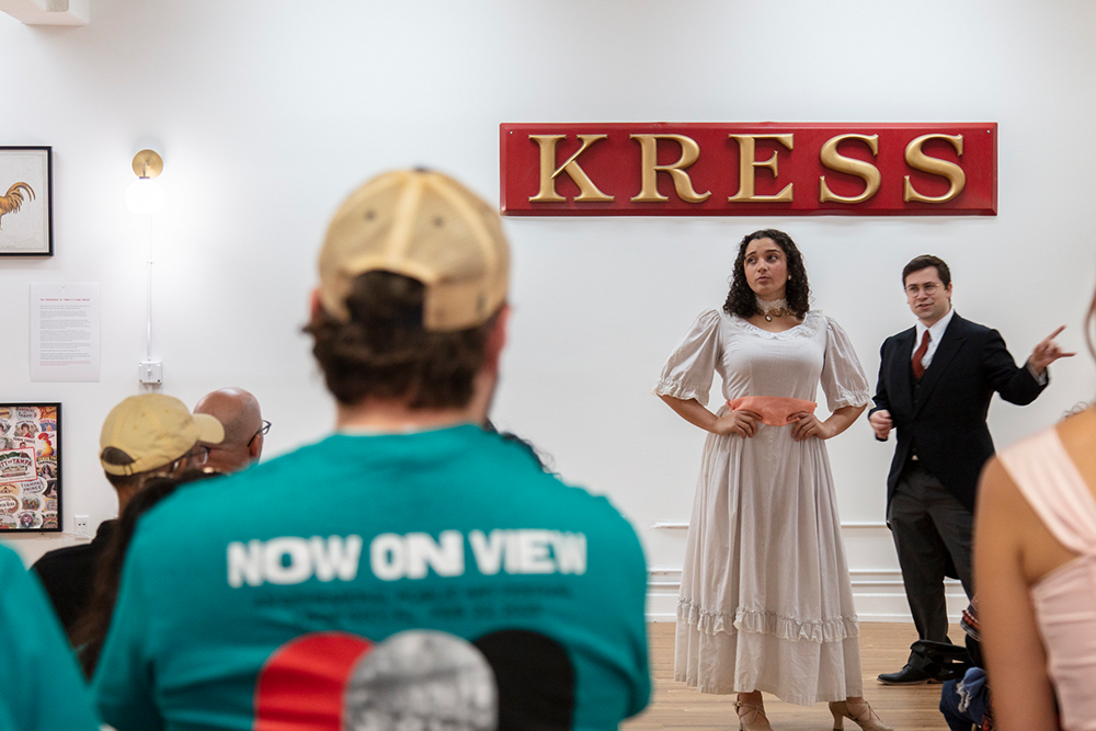 Two performers stand beneath a 'KRESS' sign in front of an audience, one in a light dress and the other in a dark suit.