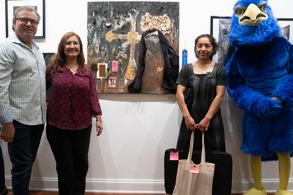 Four people stand in an art gallery, including one in a blue hawk costume, with a mixed-media artwork displayed on the wall behind them.