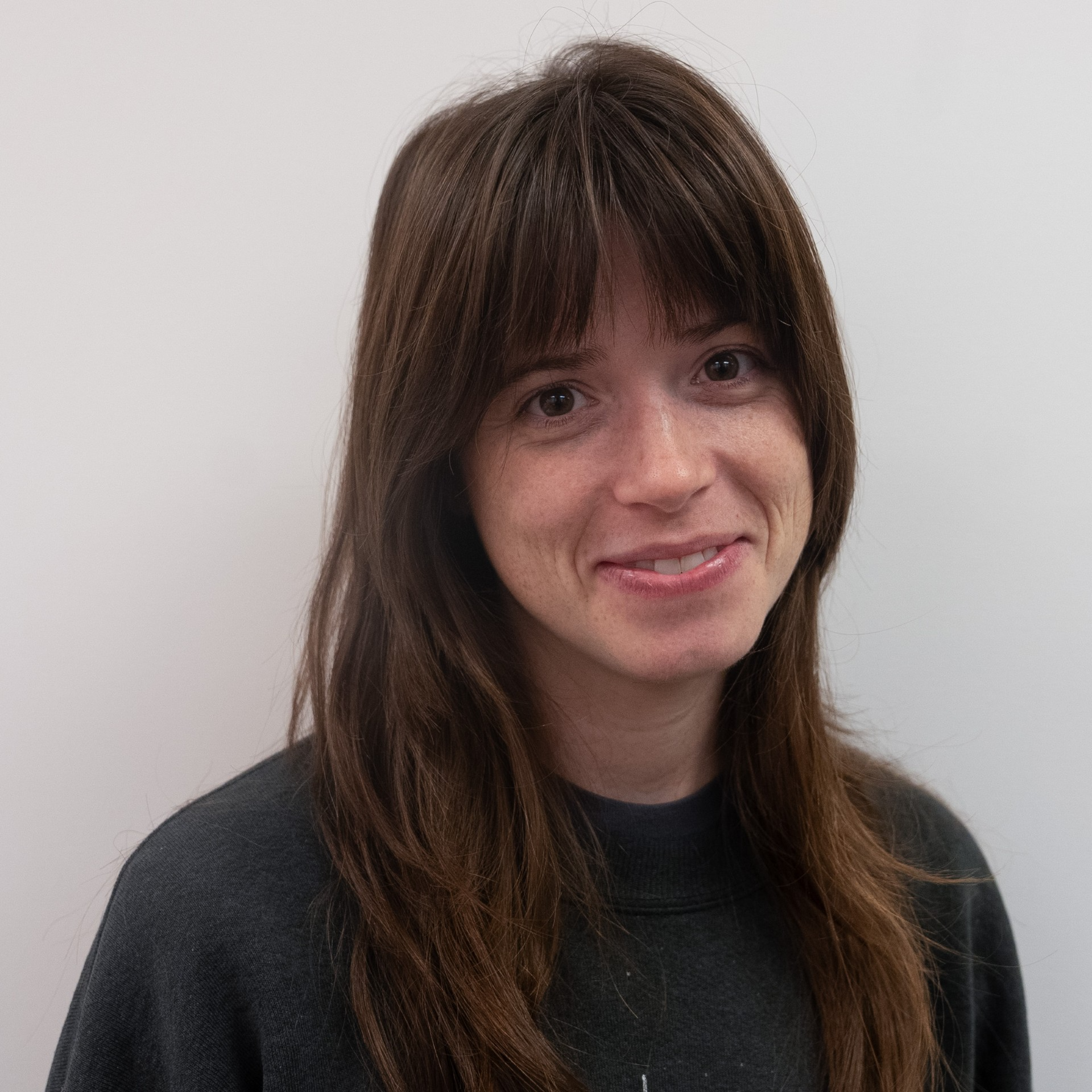 A person with long hair wearing a black sweatshirt smiles in front of a neutral background.