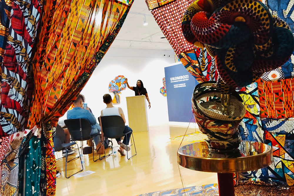 Gallery room with colorful fabric art framing a speaker at a podium, addressing a small audience.