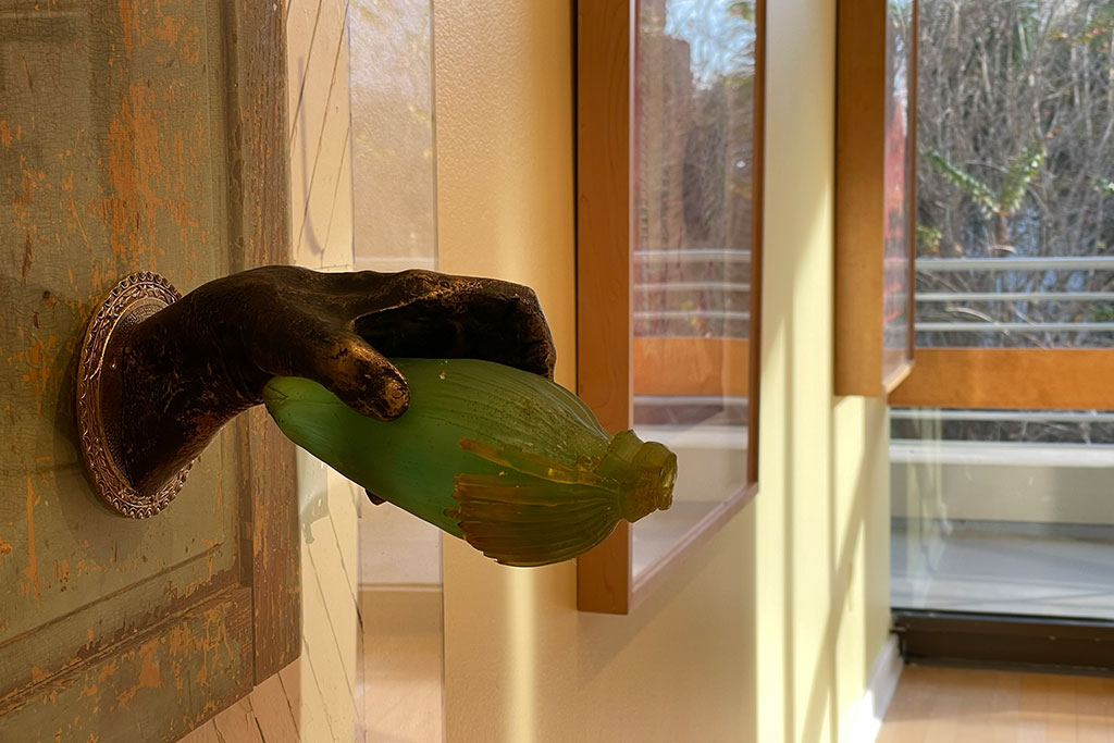 A bronze hand on a wooden wall holding a green bottle with windows in the background.