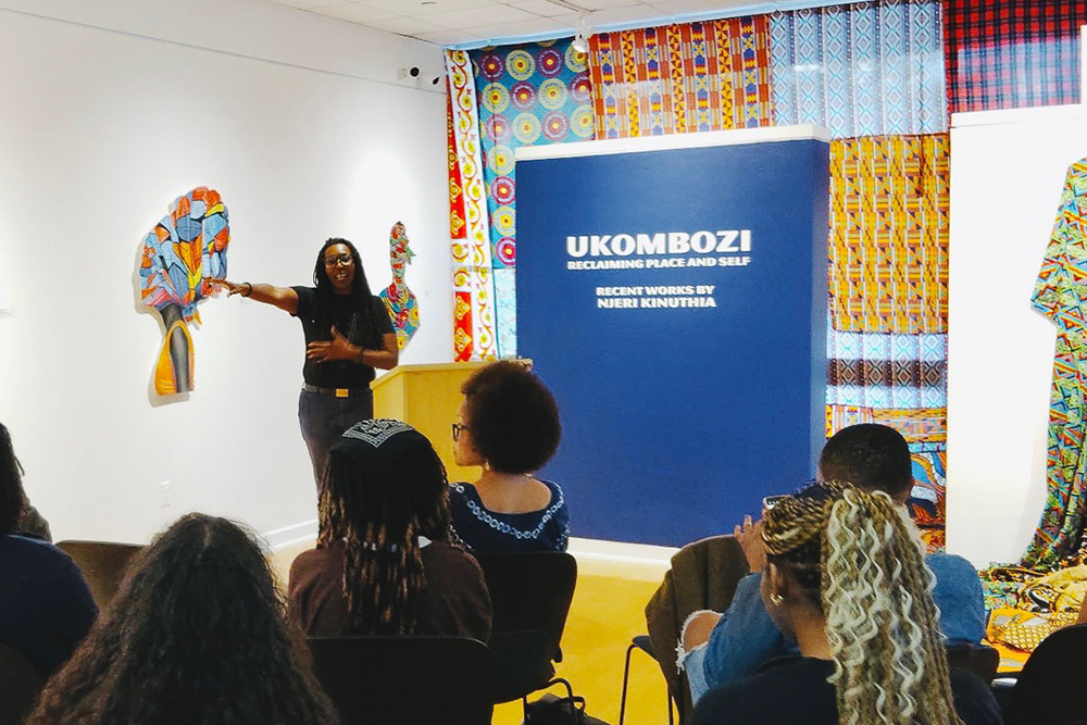 A person presents in an art gallery with colorful artwork and a display board titled "Ukombozi."