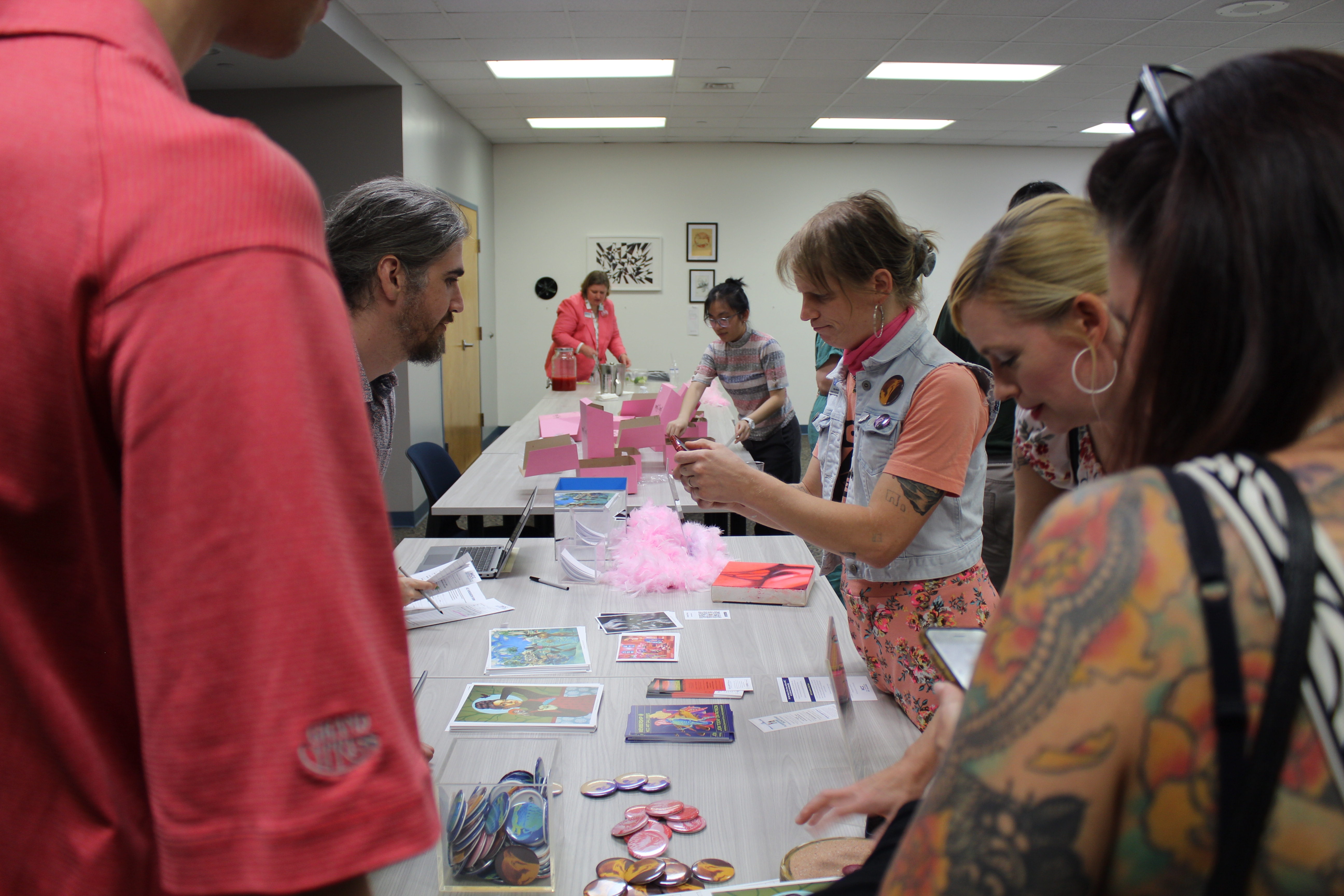 Several attendees stand around a table covered with printed materials, buttons, and event items during a Back to School Bash. The event takes place in an indoor meeting room where participants browse resources and interact with activity stations. Pink decorations and giveaway items are visible on the table, creating a festive atmosphere.