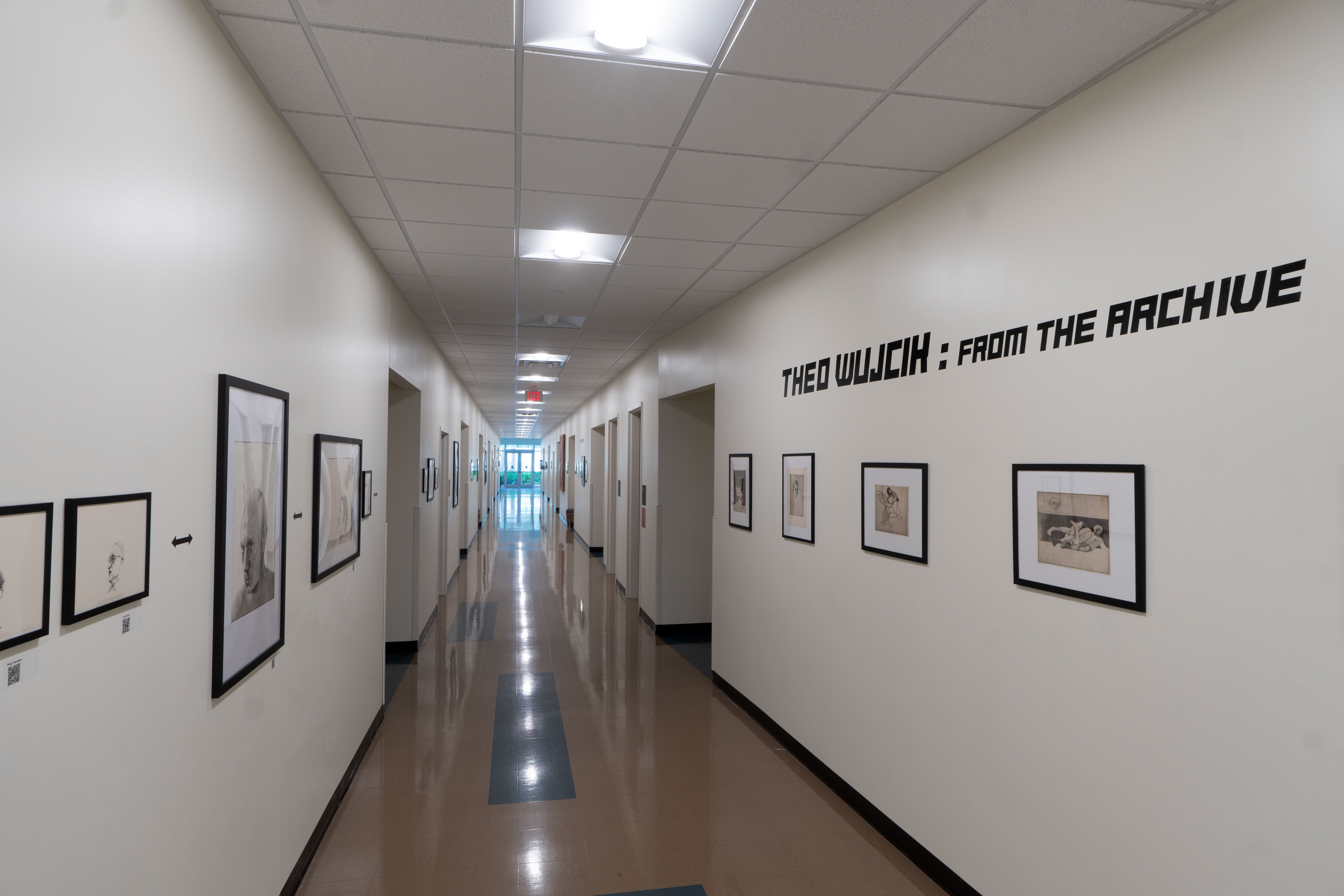 A long, brightly lit hallway is lined with framed artworks mounted on both walls. Large text reading “Theo Wujcik: From the Archive” appears on the right wall above several pieces. The exhibition extends down the corridor, creating a gallery-like display of archived artwork.