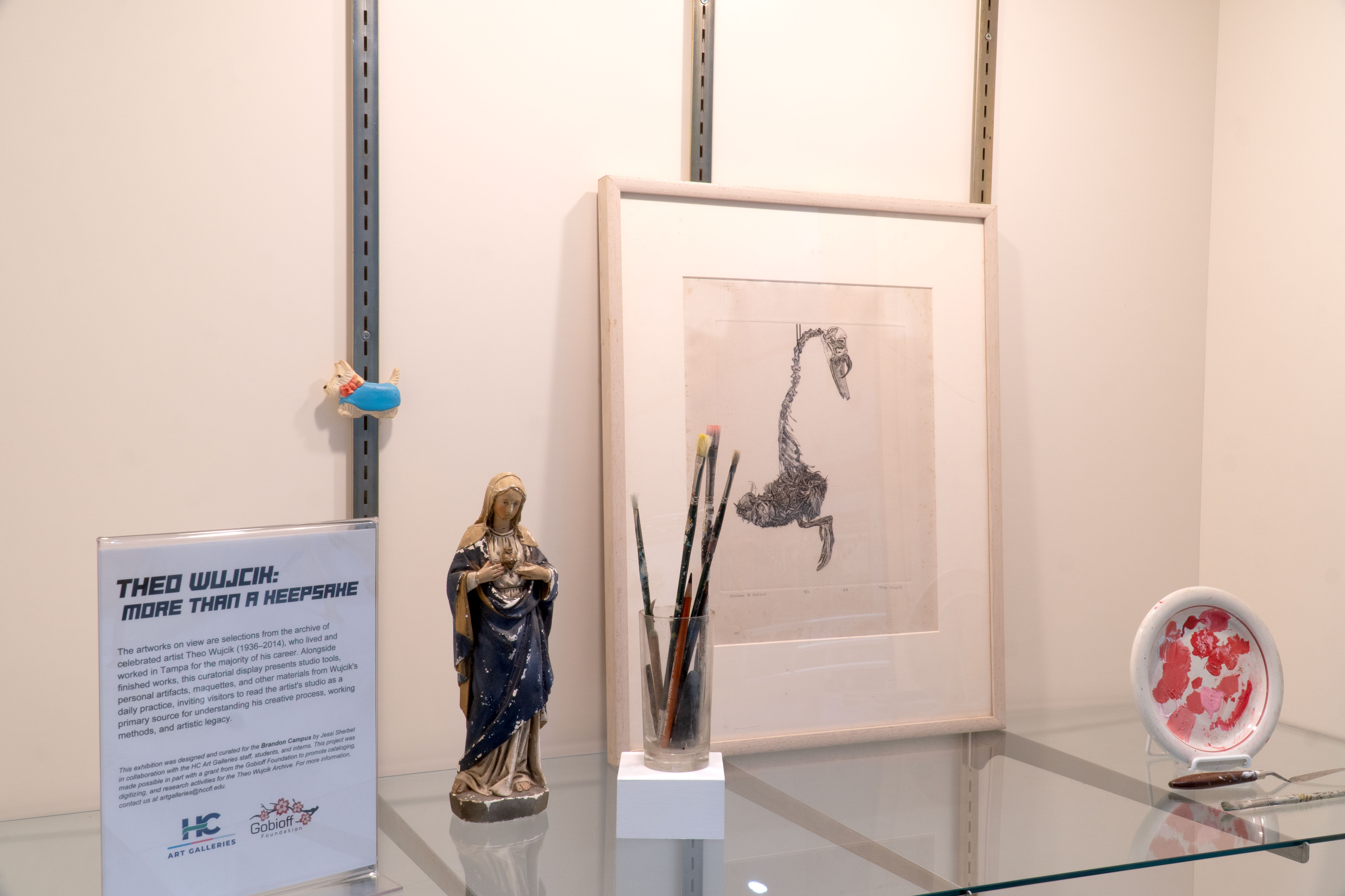 A museum-style display case presents items from the Theo Wujcik archive. The exhibit includes a framed drawing, paintbrushes in a glass container, a small figurine, and a ceramic plate with red paint marks. An exhibit sign titled “Theo Wujcik: More Than a Keepsake” provides context about the artist and the archival collection