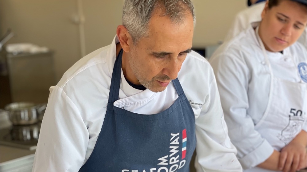 A chef in a blue apron with astudent looking on
