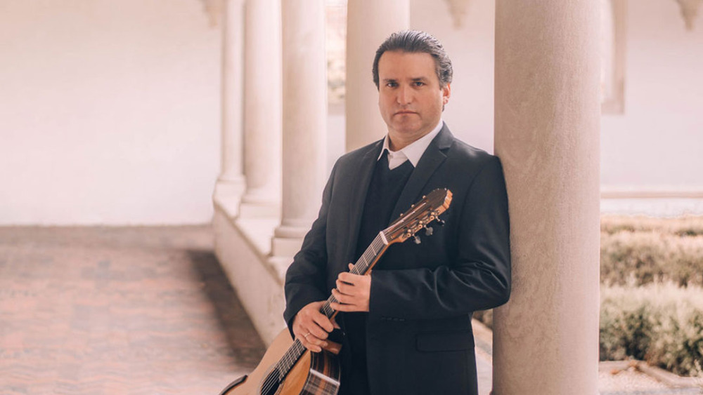 A man in a black suit holding an acoustic guitar stands next to a white column in a softly blurred architectural setting.