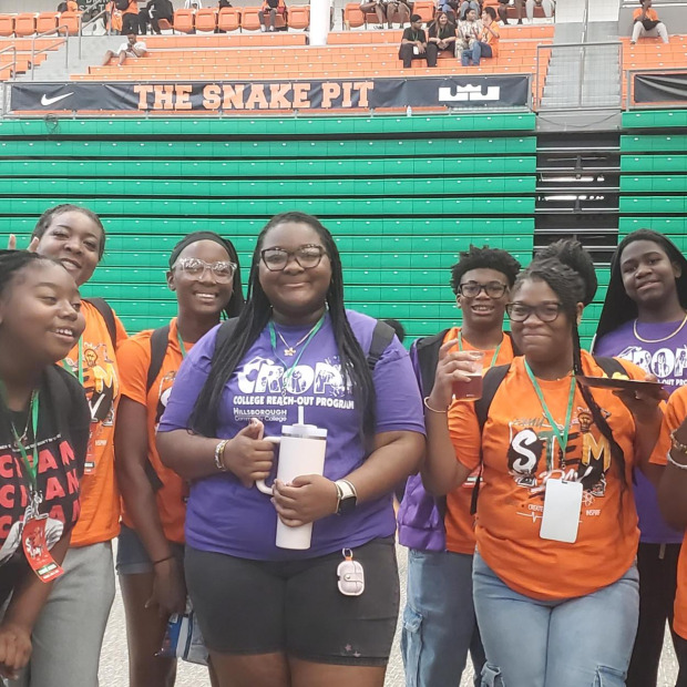 A diverse group of young people in colorful T-shirts stands together in a sports arena, smiling and posing for a photo against green bleachers.