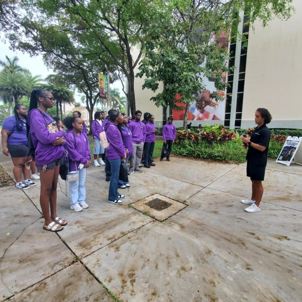 A group of individuals in purple hoodies listens to a speaker on a sidewalk outside a building with tropical landscaping and murals.