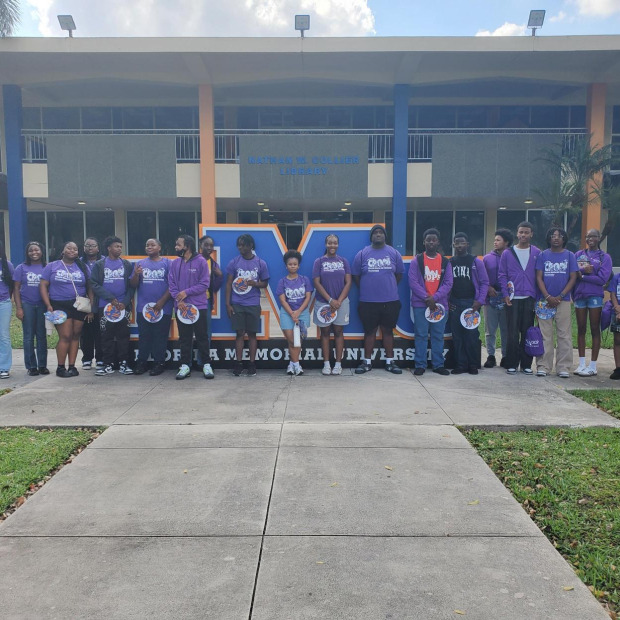 A group of approximately 20 individuals in purple shirts stands in front of a large "FMU" sign at Florida Memorial University, with a modern building and palm trees in the background.