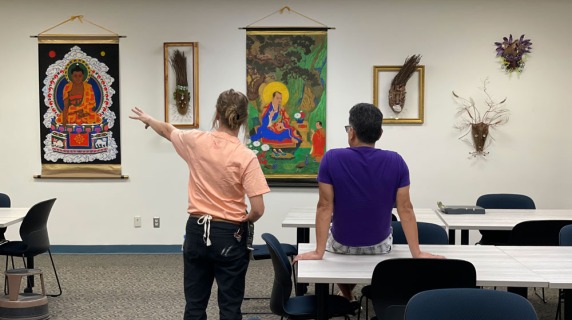 Two people in a room with Buddhist art and masks on the wall.
