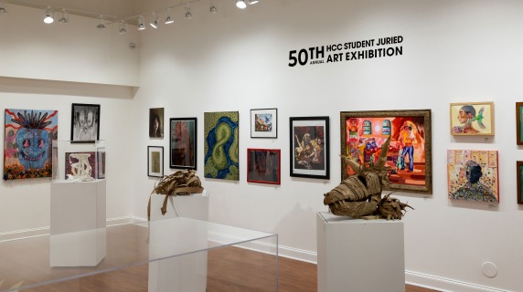 Gallery with various artworks on display, including paintings and sculptures, under bright track lighting.