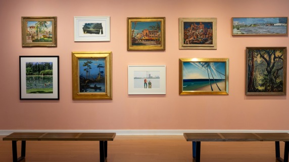 Art exhibition wall with ten paintings in various styles, framed differently, on a pink wall with two wooden benches in front.