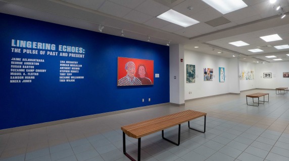 Art gallery exhibition with a blue wall featuring artist names and colorful paintings displayed.