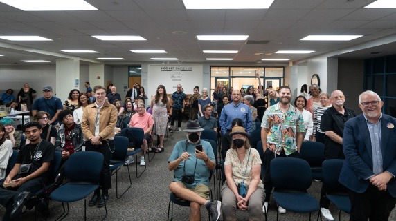 A diverse crowd gathered in a room with rows of blue chairs and a sign reading "HCC Art Galleries."
