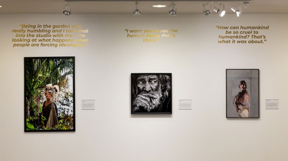A wall in an art gallery displays three framed artworks, each with a gold quote above it.