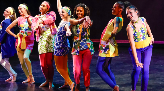 Seven dancers in colorful costumes pose energetically on a dark stage.