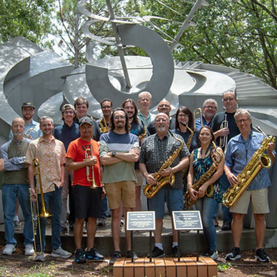 A group of people with musical instruments in front of a large silver sculpture outdoors.