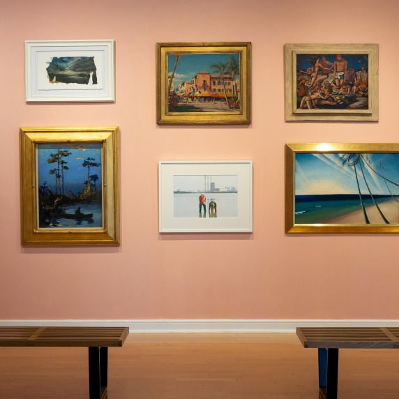 Art exhibition wall with ten paintings in various styles, framed differently, on a pink wall with two wooden benches in front.