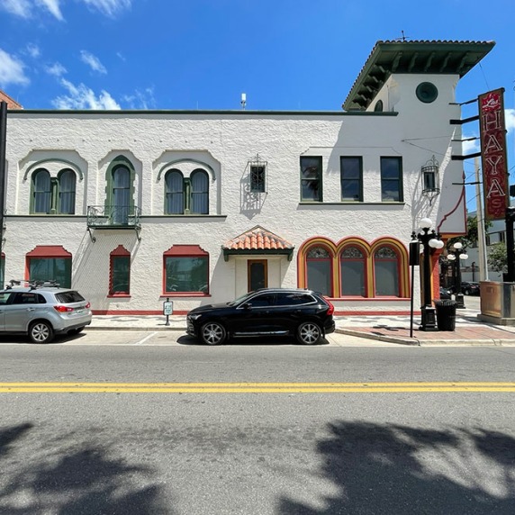A street view of a two-story white building with arched windows and cars parked in front.