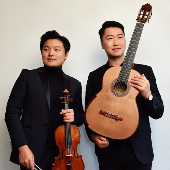 Two individuals in formal black attire, one holding a violin and the other a classical guitar.
