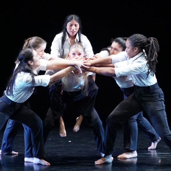 A group of six dancers in white shirts and black pants perform on a dark stage, lifting one dancer in the center.