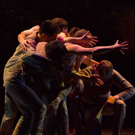 A group of dancers huddled together on a dimly lit stage with arms outstretched.