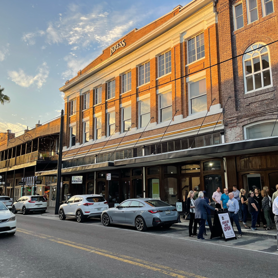 A historic brick building marked "KRESS" with people gathered below. Cars are parked along the street lined with palm trees under a partly cloudy sky.