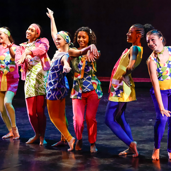 Seven dancers in colorful costumes pose energetically on a dark stage.