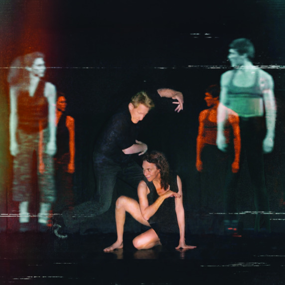 A dance performance with a woman on the floor and a man bending over her, surrounded by blurred, ghostly figures.
