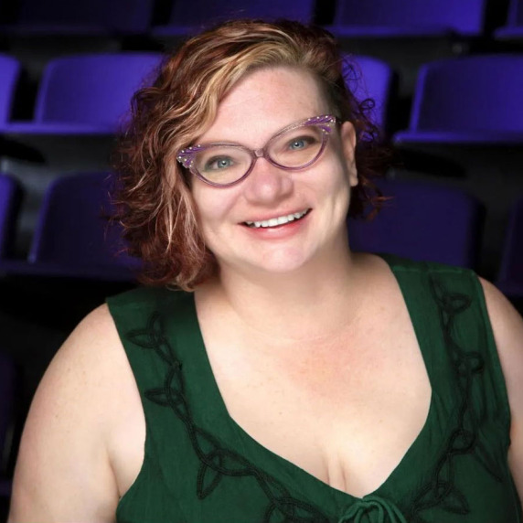 A person with purple glasses, curly red and blonde hair, and a green top smiles with purple theater seats in the background.