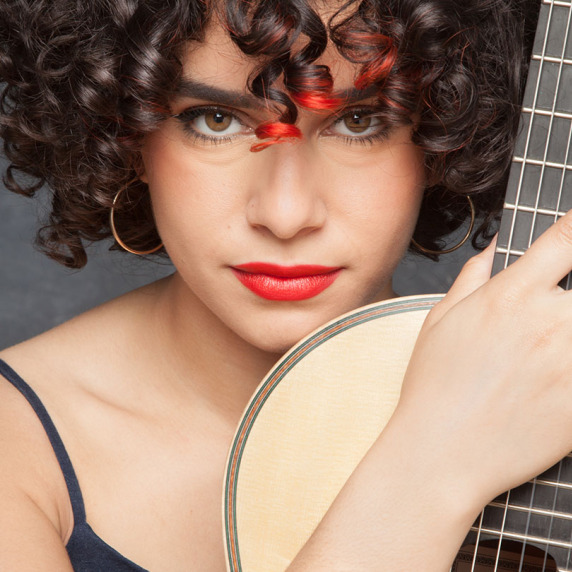 Portrait of a young woman with curly hair and red lips, holding a wooden guitar and gazing at the camera against a dark gray background.