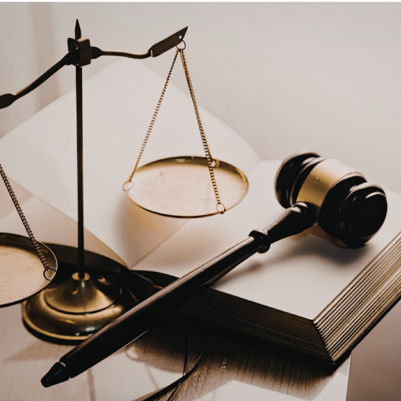 A close-up image shows a set of brass balance scales and a wooden judge’s gavel placed on an open book. The legal symbols are arranged on a polished surface with soft lighting, representing law, justice, and the judicial process.