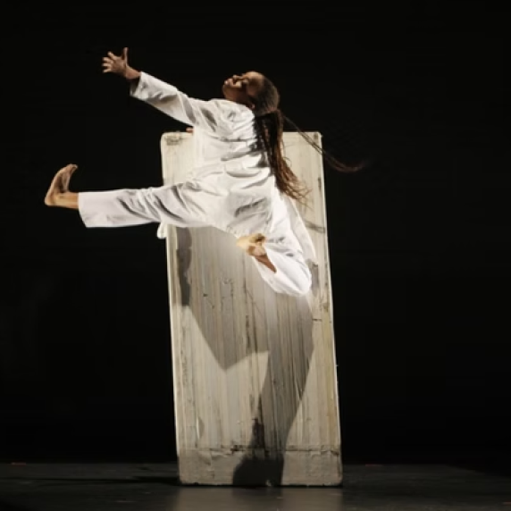 Dancer in a white costume leaps across a stage in front of a tall rectangular set piece, captured midair with arms and legs extended.