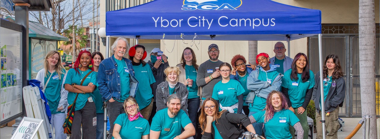 Group of people under a blue tent labeled "Ybor City Campus", with palm trees and a building behind.