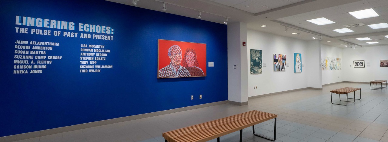 Art gallery exhibition with a blue wall featuring artist names and colorful paintings displayed.
