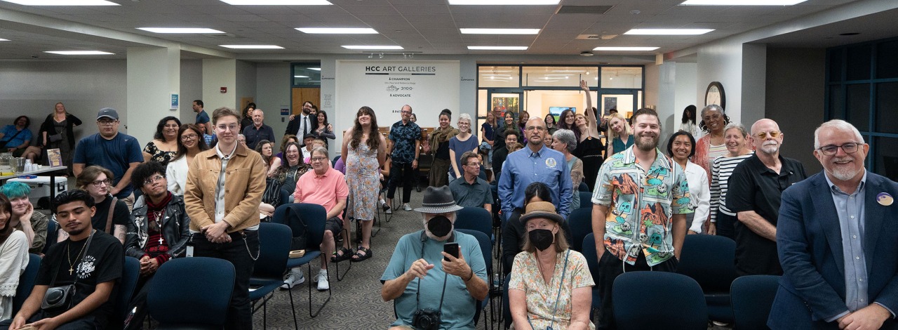 A diverse crowd gathered in a room with rows of blue chairs and a sign reading "HCC Art Galleries."