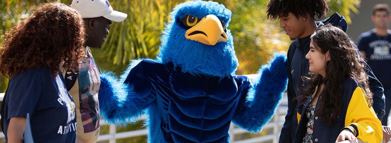 Mascot in a blue bird costume interacting with young people on a sunny walkway.