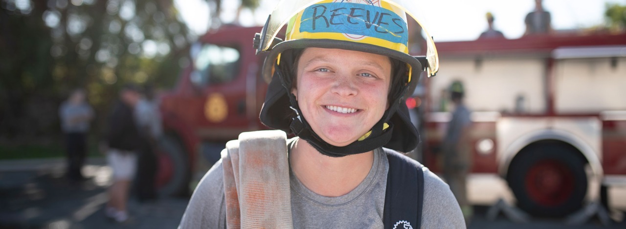 Firefighter smiling with hose over shoulder, red fire truck in background.