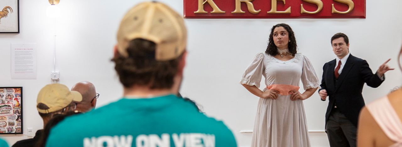 Two performers stand beneath a 'KRESS' sign in front of an audience, one in a light dress and the other in a dark suit.