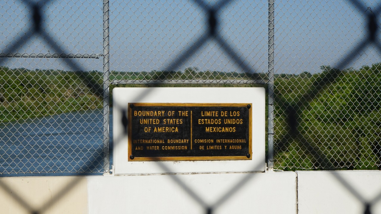 A chainlink fence with a sign that reads "boundary of the United States of America" seen through another chainlink fence