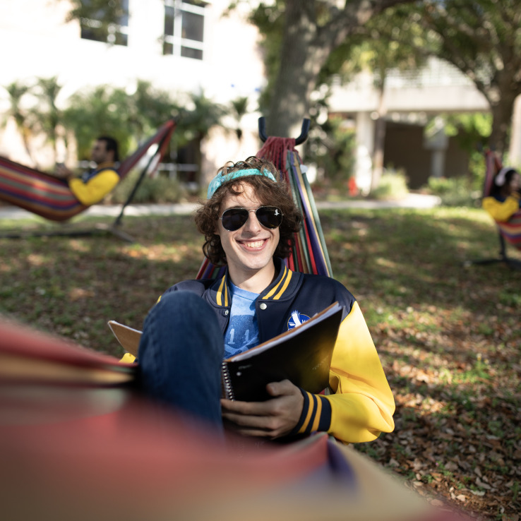 Male student wearing sunglasses and smiling while lounging in a hammock.